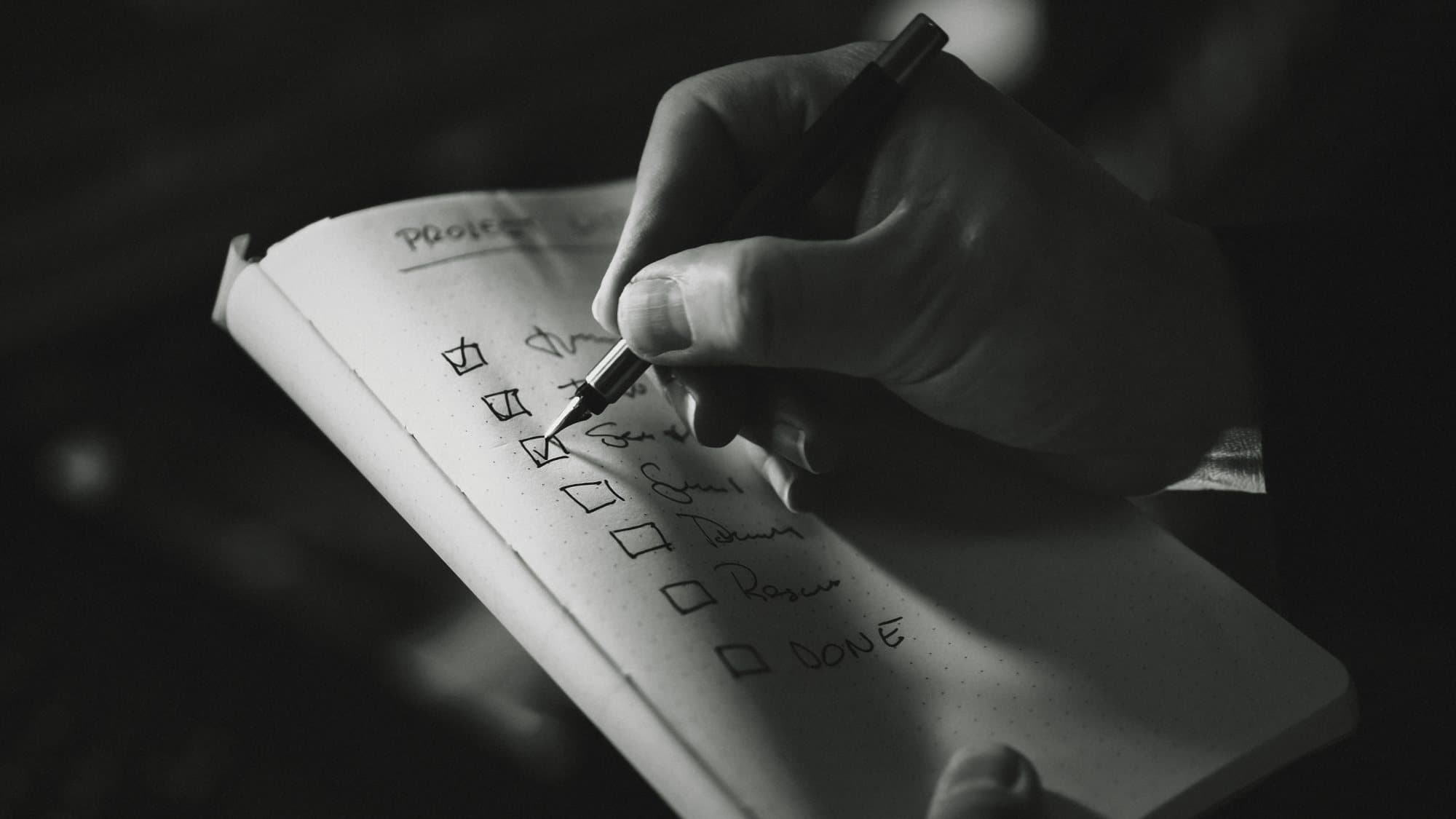 A hand ticking boxes on a handwritten checklist in a notebook, in black and white.