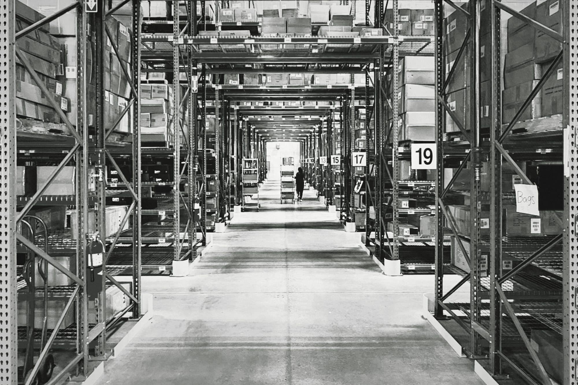 A long aisle between tall warehouse shelving, with a person pushing a cart in the distance, in black and white.